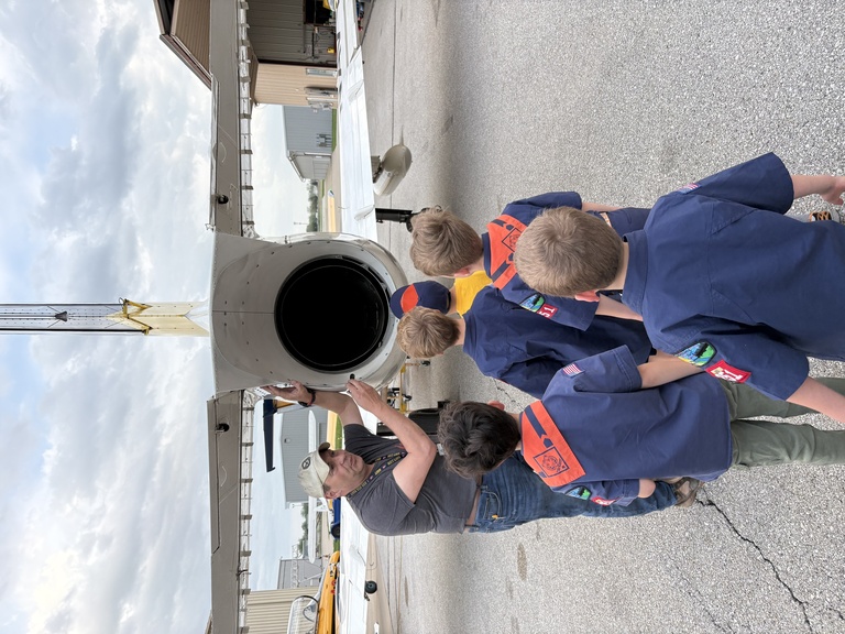 Children in blue uniform shirts with red handkerchief look on as man with cap and short sleeve blue shirt points at exhaust of jet.