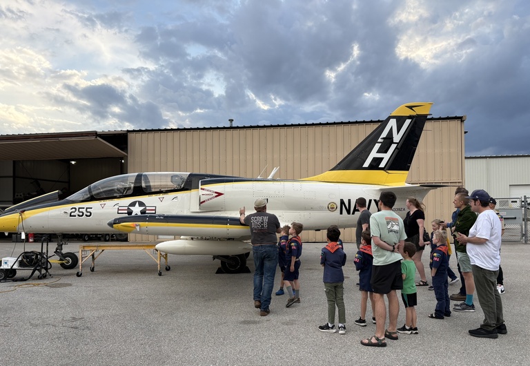 Adults and children stand on pavement looking at white jet with black and yellow accents