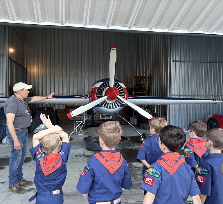 Children in blue uniform shirts with red handkerchief in the foreground with airplane with propeller facing forward in the background and man with blue short sleeve shirt and cap stands to the left