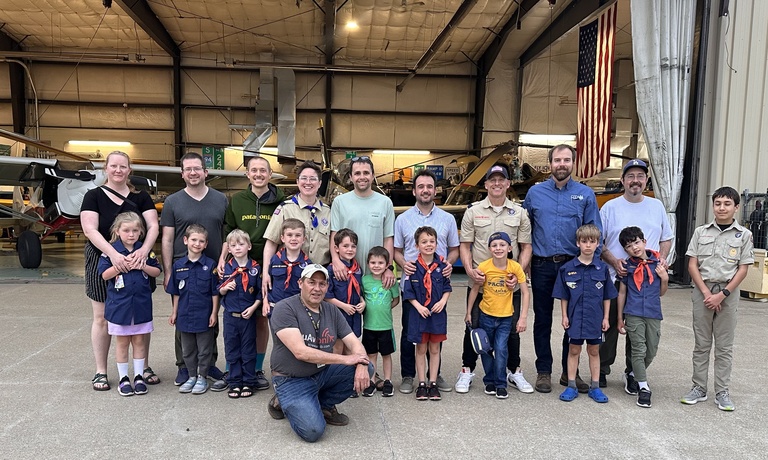 A group photo with a row of nine adults standing behind a row of 11 children with man in cap and blue shirt crouched in front toward the right, all in front of a hangar