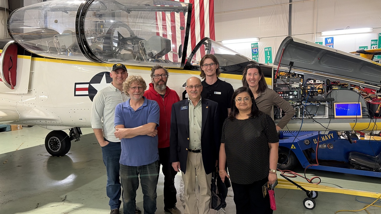 Seven people stand in front of jet in hangar with American flag hanging vertically in background