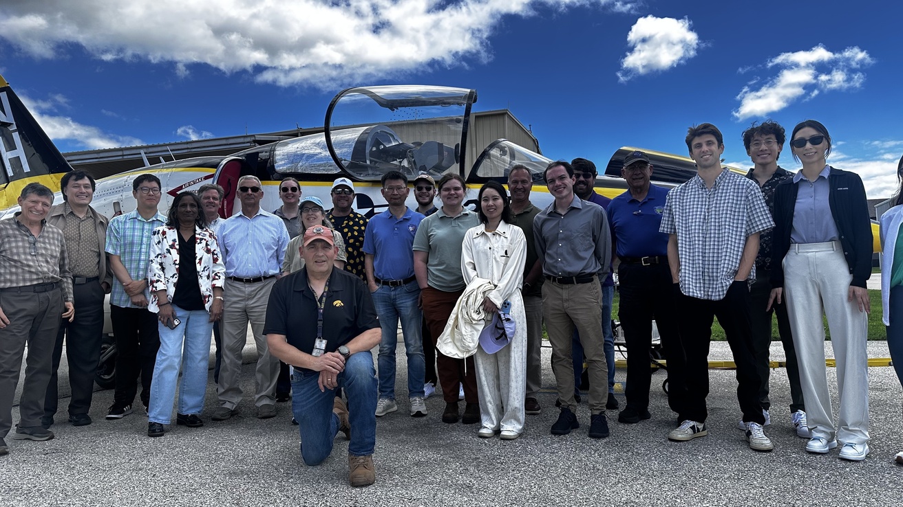 Group of people stand while one kneels, all outside in front of a white jet.